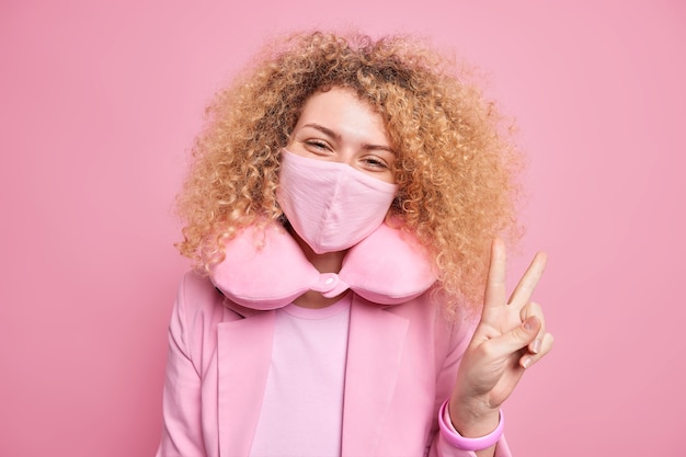 Pleased carefree young woman wears face mask to protect from coronavirus got used to quarantine measurements makes peace sign dressed in elegant clothes uses neck pillow for feeling comfort.