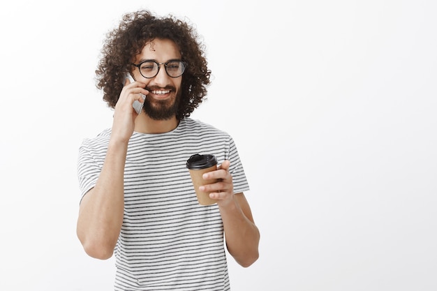 Tシャツとメガネのカジュアルなボーイフレンドを喜ばせ、紙コップからコーヒーを飲み、わずかな笑顔でスマートフォンで話し、よそ見