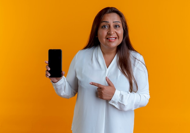 Pleased casual caucasian middle-aged woman holding and points at phone 