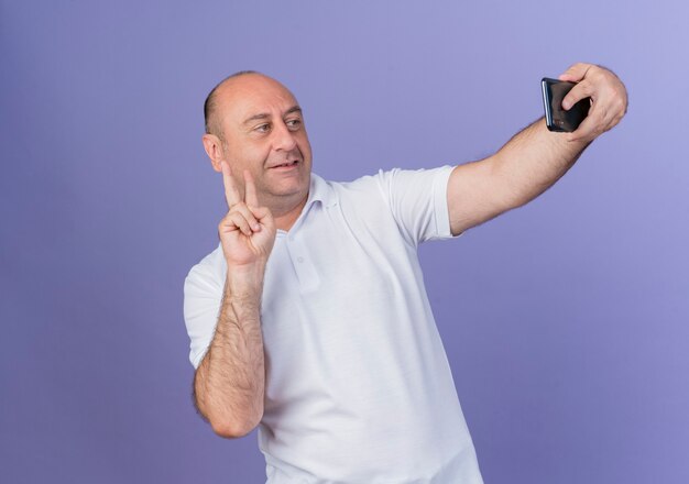 Pleased casual mature businessman doing peace sign and taking selfie isolated on purple background with copy space