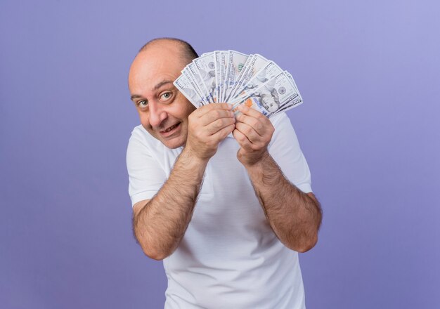 Довольный случайный зрелый бизнесмен держит деньги и смотрит в камеру из-за денег, изолированных на фиолетовом фоне с копией пространства