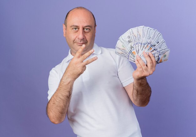 Довольный случайный зрелый бизнесмен, держащий деньги, смотрящий в сторону и показывающий три рукой, изолированной на фиолетовом фоне с копией пространства
