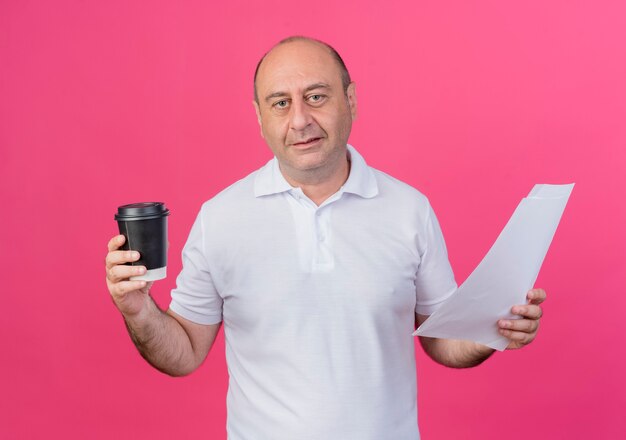 Доволен случайный зрелый бизнесмен, держащий пластиковую кофейную чашку и документы, изолированные на розовом фоне