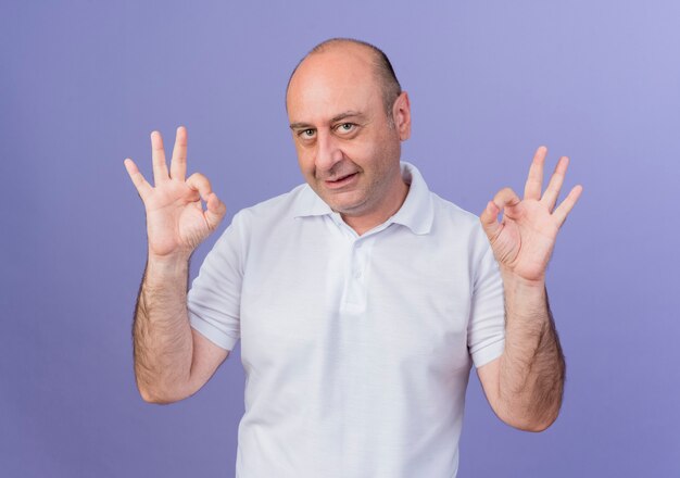 Pleased casual mature businessman looking at camera and doing ok signs isolated on purple background