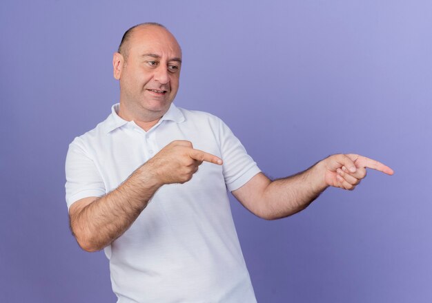 Pleased casual mature businessman looking and pointing at side isolated on purple background
