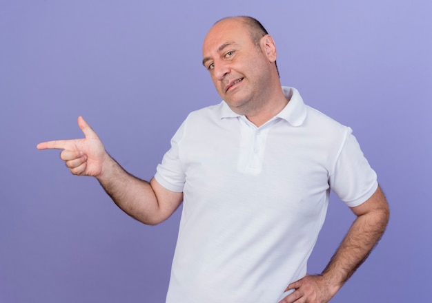 Pleased casual mature businessman putting hand on waist and doing pistol gesture at side isolated on purple background