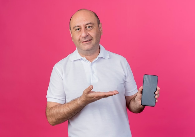 Pleased casual mature businessman showing mobile phone and pointing at it with hand isolated on pink background