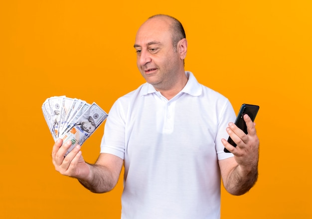 Pleased casual mature man holding phone and looking at money in his hand isolated on yellow backgound