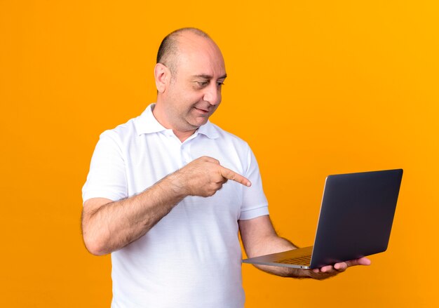 Pleased casual mature man holding and points at laptop isolated on yellow backgound