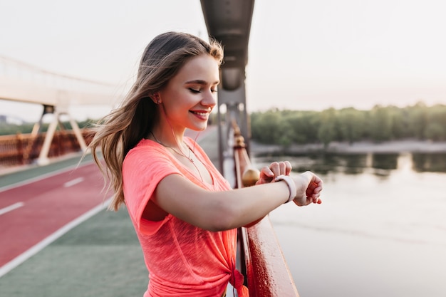 Pleased caucasian girl in casual t-shirt using fitness bracelet. Outdoor shot of stunning lady smiling after training.