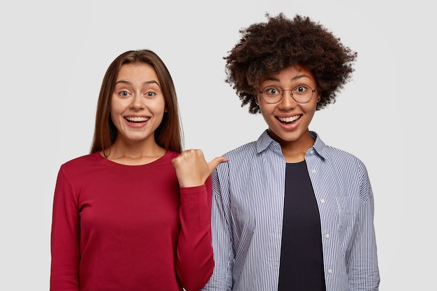 pleased Caucasian woman with joyful expression, points at her African American friend