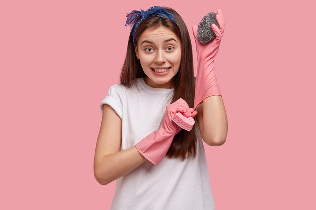 Pleased Caucasian young woman puts on rubber gloves, carries two sponges, ready to wash dishes