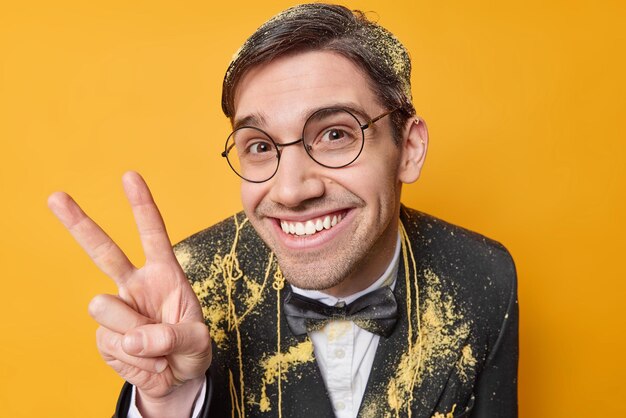 Pleased cheerful adult man smiles gladfully being in good mood wears round spectacles and festive black formal suit enjoys celebration isolated over yellow studio wall Hey stay always positive