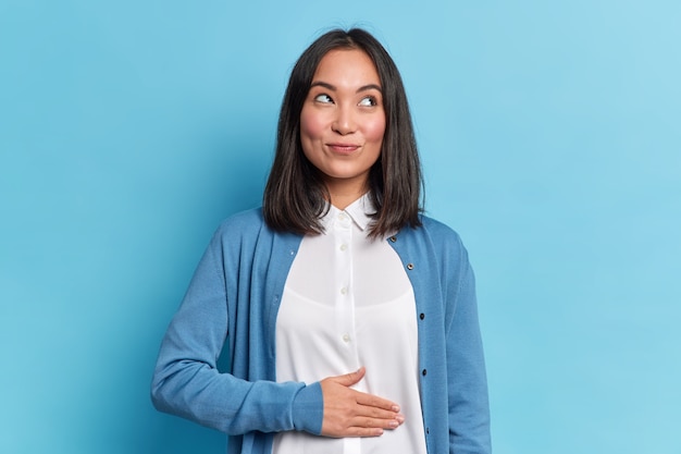 Foto gratuita la donna asiatica allegra e contenta tiene la mano sulla pancia si sente piena dopo una cena deliziosa vestita casualmente sta pensierosa