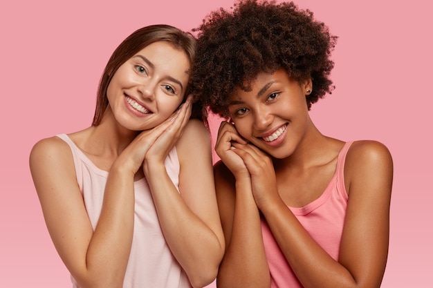 Pleased cheerful dark skinned young woman stands closely to her best Caucasian friend, model against pink wall, express positive emotions and feelings. Multicultural companions pose indoor