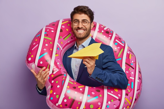 Pleased cheerful male director returns at work after summer holiday, throws paper plane, stands in inflated swimring, smiles positively