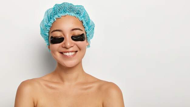 Pleased cheerful young woman has beauty undereye patch treatment, closes eyes from satisfaction, wears waterproof headgear, stands with naked body