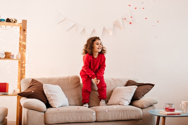 ソファでジャンプして喜んでいる子供。ソファの上に立っているかわいいプレティーンの女の子の屋内ショット。