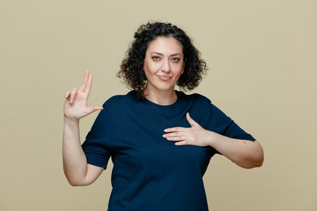 Pleased and confident middleaged woman wearing tshirt looking at camera showing promise gesture isolated on olive green background