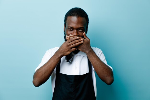 Pleased covered mouth with hand holding hair clippers young african american barber in uniform isolated on blue background