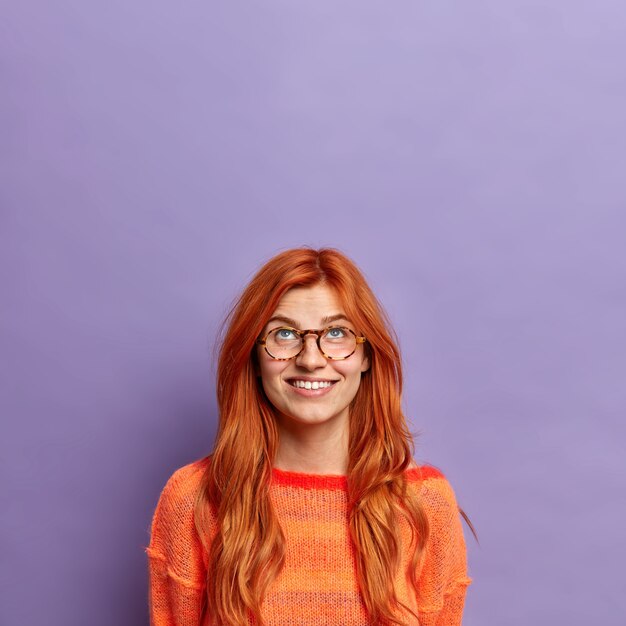 광활한 미소로 위에 집중된 호기심 많은 빨간 머리 유럽 여성은 캐주얼 한 옷을 입은 광학 안경을 착용합니다.