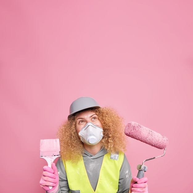 Free photo pleased curly female builder or laborer wears helmet and protective respirator unform holds paint roller and brush focused above