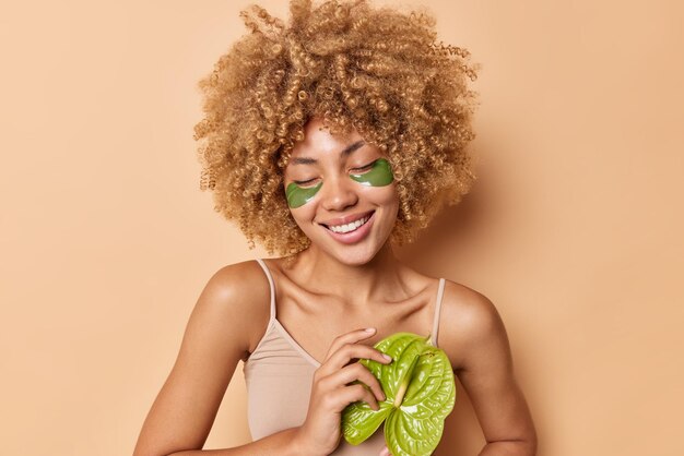 Pleased curly haired with natural makeup and green collagen patches on fresh facial skin holds calla wears casual t shirt isolated over beige background. Natural cosmetics and beauty concept