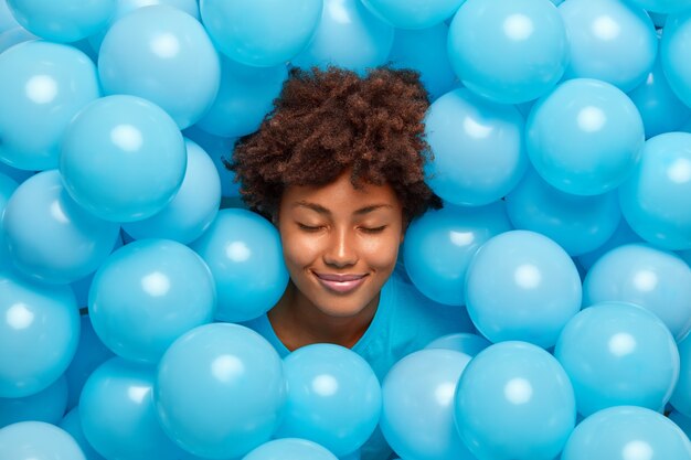 Pleased curly haired woman closes eyes surrounded by many blue inflated balloons has festive mood has fun on party feels very happy