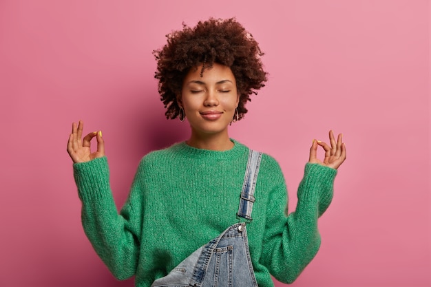 Free photo pleased curly haired woman tries to calm down, unites with nature, raises hands and shows zen gesture, meditates or does yoga indoor, closes eyes, enjoys peaceful atmosphere for good relaxation