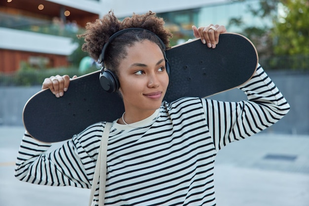 Pleased curly millennnial girl holds skateboard enjoys favorite activity listens audio track in headphones wears casual striped jumper stands outdoors against blurred background