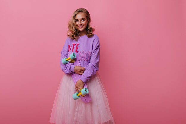 Pleased curly woman holding pink longboard and smiling. Winsome female model in lush skirt posing on pastel with sincere emotions.