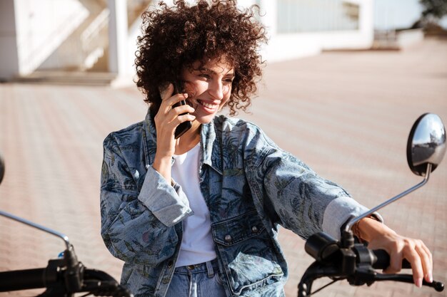 Довольная кудрявая женщина сидит на современном мотоцикле на свежем воздухе и разговаривает по смартфону