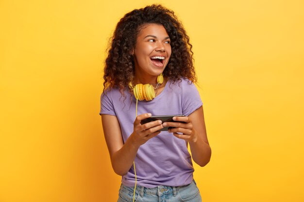 Pleased curly woman with Afro haircut, holds smartphone horizontally, plays games online