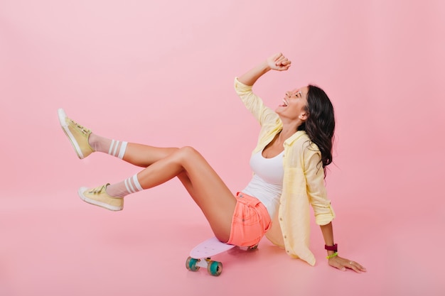 Pleased dark-haired girl in cute shorts and colorful socks playfully posing on skateboard. Indoor portrait of tanned young woman in bright accessories sitting and laughing.