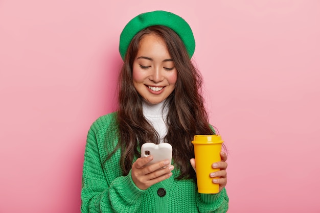 Pleased dark haired girl focused in cell phone, glad to get message of invitation for party, surfes social networks on modern gadget, checks newsfeed, wears green fashionable clothes, drinks coffee