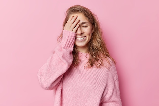 Pleased dark haired woman makes face palm tries to hide herself keeps eyes shut smiles gently laughs at something wears casual jumper isolated over pink background. Monochrome shot. Blind concept.