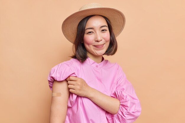 루즈한 뺨을 가진 기쁘게 검은 머리 젊은 아시아 여성은 백신 접종 후 회반죽 팔을 보여주고 베이지색 벽 위에 격리된 분홍색 블라우스와 페도라
