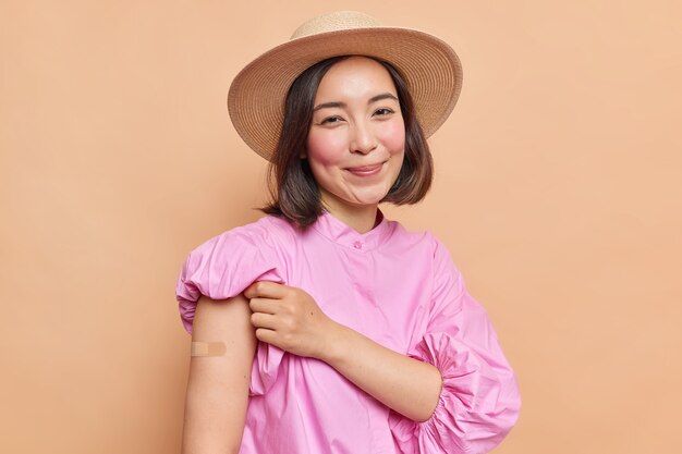 Pleased dark haired young Asian lady with rouge cheeks shows plastered arm after vaccination weras fedora and pink blouse isolated over beige wall