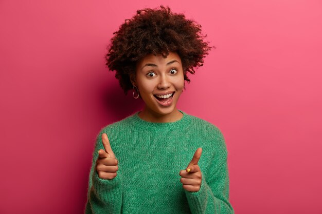 Pleased dark skinned female makes finger gun pistol, indicates at you, expresses choice, smiles pleasantly, wears green jumper, isolated on pink wall, smiles upbeat, congratulates colleague