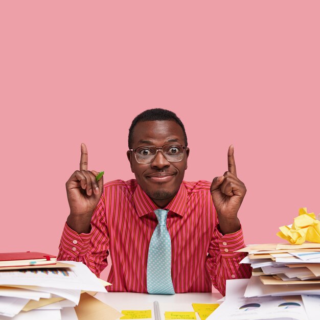 Pleased dark skinned man searches creative soultion for improving project, makes decision, has pile of documents on desk