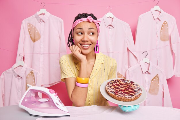 Pleased dark skinned multitasking housewife bakes pie and irons clothes for family wears headband bracelets poses near ironing board smiles happily poses against pink wall