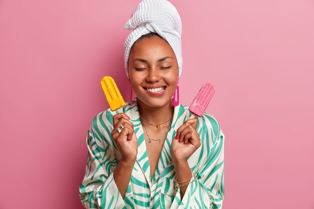 Pleased dark skinned woman bites lips, holds tasty appetizing ice cream, imagines pleasant taste of frozen dessert, wears wrapped towel on head, dressed in gown, 