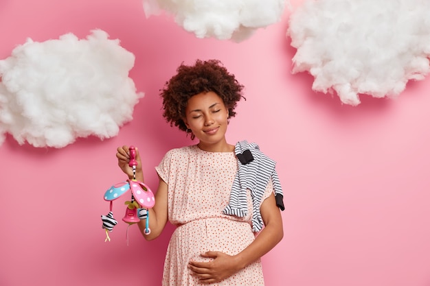 Foto gratuita lieta donna dalla pelle scura che accarezza la sua pancia incinta sente il suo bambino appena nato chiude gli occhi e sta calmo gode di gravidanza porta oggetti per bambini pone