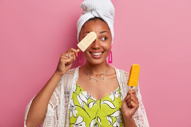 Pleased dark skinned woman covers eye with delicious ice cream on eye smiles broadly