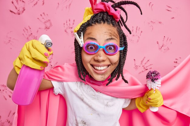 Pleased dark skinned woman dressed like superhero sprays detergent holds toilet brush wears goggles cape appreciates cleanliness smiles gladfully isolated over pink wall