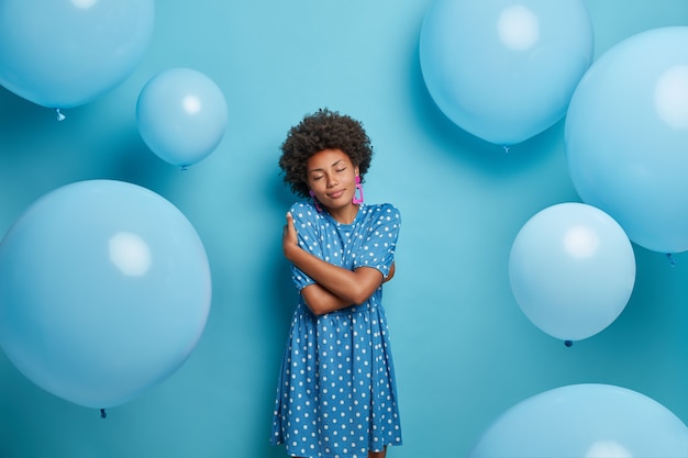 Pleased dark skinned woman embraces herself and closes eyes with pleasure, poses with blue balloons