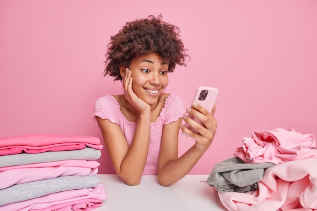 곱슬머리를 한 어두운 피부의 여성이 스마트폰을 통해 뉴스피드를 통해 분홍색 벽에 격리된 세탁 후 접힌 옷 주위에 세탁물 더미가 있는 테이블에서 포즈를 취합니다. 가구 개념