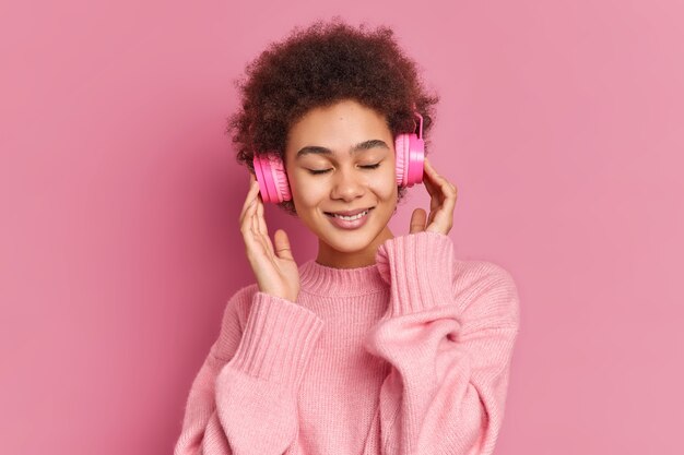 Pleased dark skinned young woman enjoys listening pleasant melody keeps hands on stereo headphones closes eyes wears casual jumper 