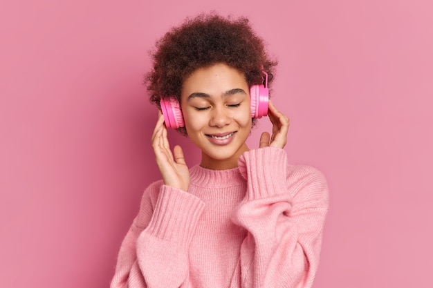 Pleased dark skinned young woman enjoys listening pleasant melody keeps hands on stereo headphones closes eyes wears casual jumper 