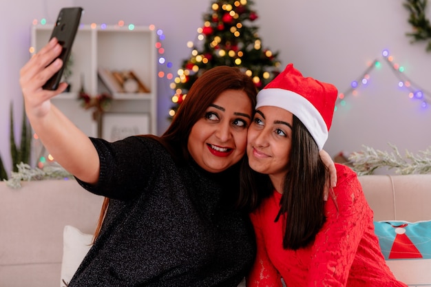 Pleased daughter with santa hat and mother take selfie looking at phone sitting on couch enjoying christmas time at home